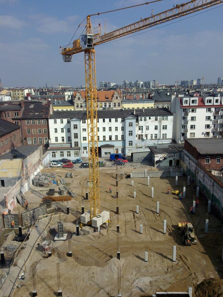 Widok z góry na plac budowy w centrum miasta z wysokim, żółtym dźwigiem wieżowym, widoczne fundamenty pod budynek i pracujący robotnicy.