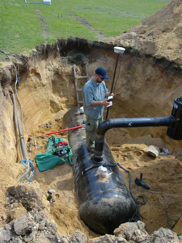 Instalacja zbiornika w wykopie, pracownik z urządzeniem pomiarowym GPS sprawdza poziom, widoczne narzędzia i rury.