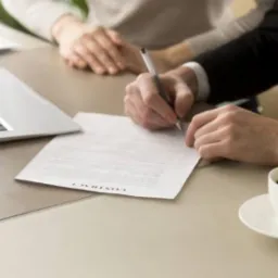 Mężczyzna w garniturze podpisuje umowę przy biurku, obok laptop, kawa i dłoń drugiej osoby.