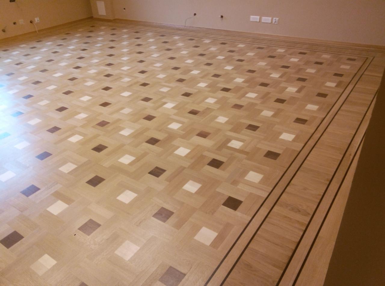 Jasny parkiet ułożony we wzór szachownicy z ciemnymi i białymi akcentami, obramowany dekoracyjną listwą przypodłogową z ciemnymi paskami.