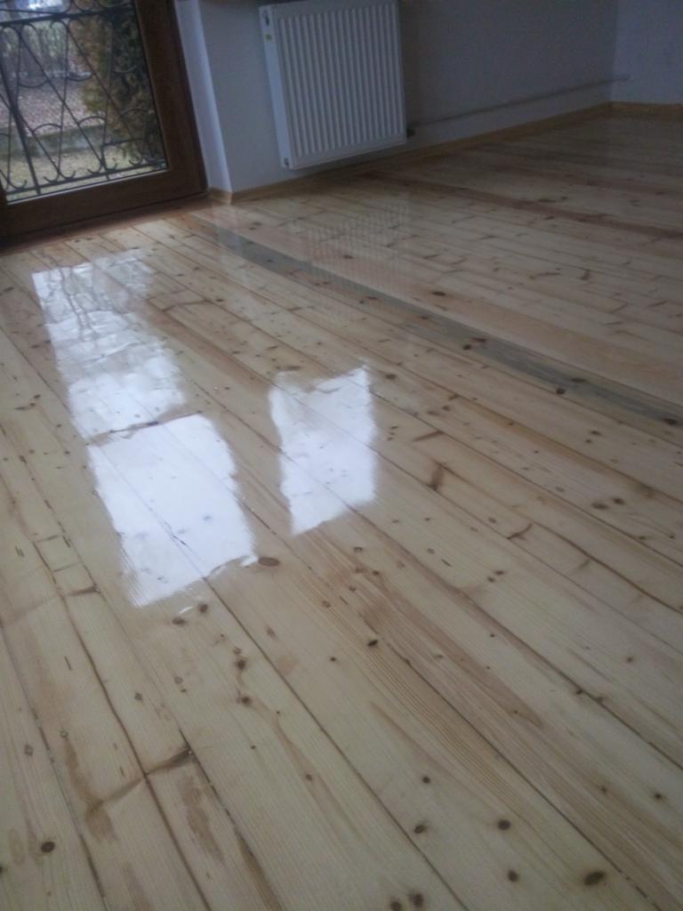 Lakierowana, odnowiona podłoga z desek sosnowych w pokoju z oknem i grzejnikiem, widoczne refleksy światła.
