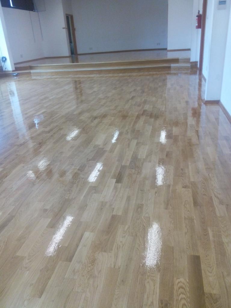 Błyszcząca, odnowiona podłoga parkietowa z wyraźnym rysunkiem drewna, po zabiegu renowacji, z widocznymi refleksami światła, w przestronnym pomieszczeniu z podwyższeniem.