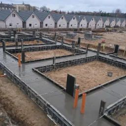 Zalane betonem fundamenty pod budowę domów jednorodzinnych w zabudowie szeregowej, widoczne szalunki, wystające rury instalacyjne i pracownicy w oddali.