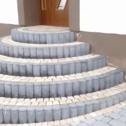 P&oacute;łokrągłe schody wejściowe, wyłożone szarą kostką obrzeżową i jasną kostką wewnątrz, prowadzące do brązowych drzwi wejściowych.