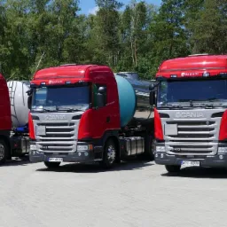 Trzy czerwone ciężarówki Scania z cysternami, ustawione równolegle na parkingu, widok z przodu, w tle drzewa.