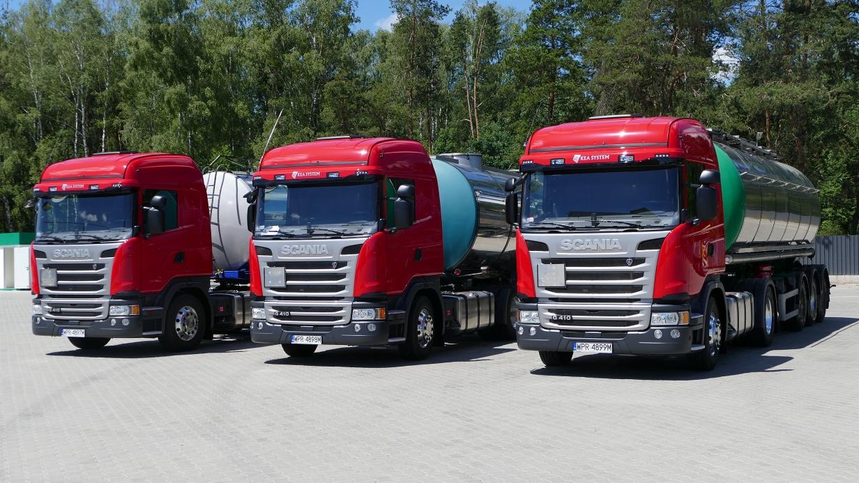 Trzy czerwone ciężarówki Scania z cysternami, ustawione równolegle na parkingu, widok z przodu, w tle drzewa.