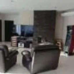 Jasny salon z czarnymi skórzanymi fotelami, ciemną ścianą z kamienia dekoracyjnego za telewizorem i lśniącą podłogą. Widoczne głośniki, szafka RTV i wysoki, wąski element dekoracyjny w kolorze...