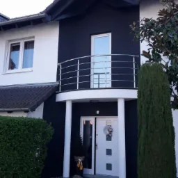 Dwukondygnacyjny dom z ciemną elewacją, balkonem z metalową balustradą i dekoracyjnym wieńcem na białych drzwiach wejściowych.