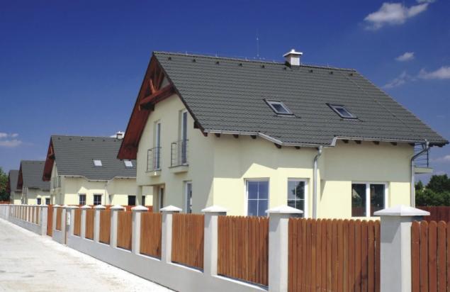 Rząd domów jednorodzinnych z jasną elewacją, szarą dachówką i drewnianym ogrodzeniem w słoneczny dzień.