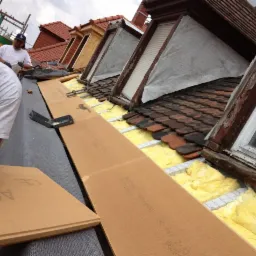 Montaż izolacji termicznej na dachu z wykorzystaniem płyt izolacyjnych i wełny mineralnej, widoczne okna dachowe i dachówka ceramiczna.