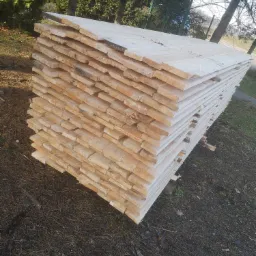 Stos świeżo przetartego drewna ułożony na zewnątrz, widoczne słoje i jasny kolor drewna, tło stanowią drzewa iglaste.