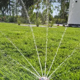 Automatyczny zraszacz rotacyjny nawadnia soczysty trawnik na tle drzew i białego budynku w słoneczny dzień.