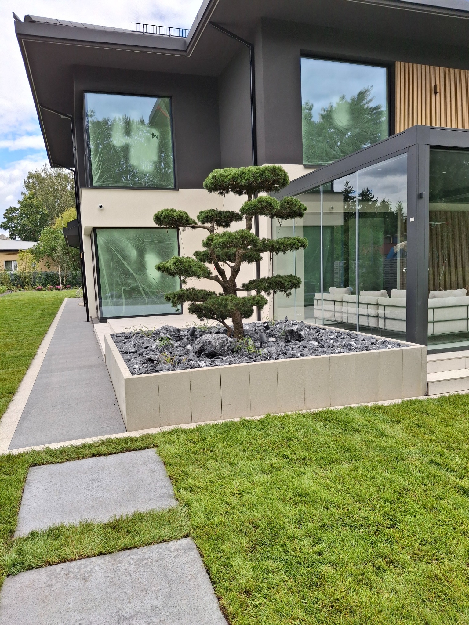 Nowoczesny ogród z bonsai w geometrycznej donicy z kamieniami, na tle domu z dużymi oknami i zadbanym trawnikiem.