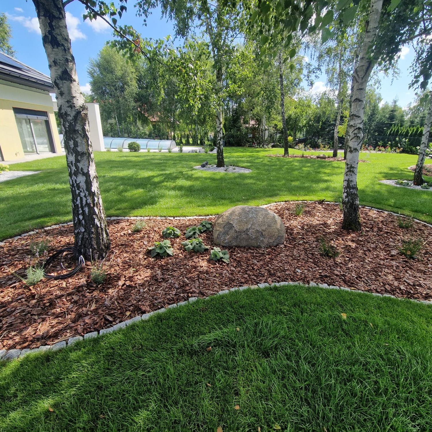 Zadbany ogród z równo przystrzyżonym trawnikiem, ozdobnym kamieniem i korą wokół drzew, obrzeża z kostki. W tle budynek z basenem.