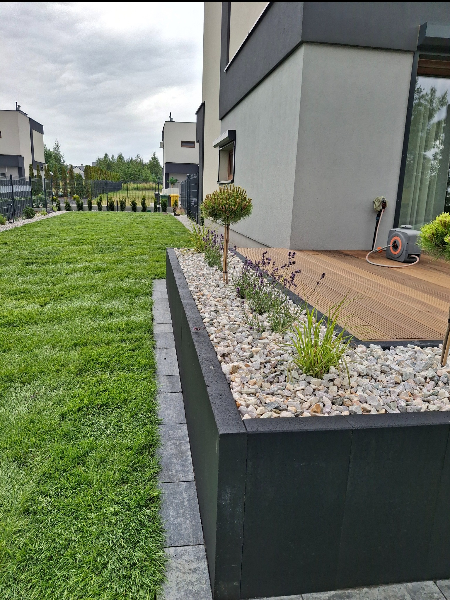 Nowoczesny ogród z równo przystrzyżonym trawnikiem, rabatą z kamieniami i roślinami, oraz drewnianym tarasem przy budynku w minimalistycznym stylu.