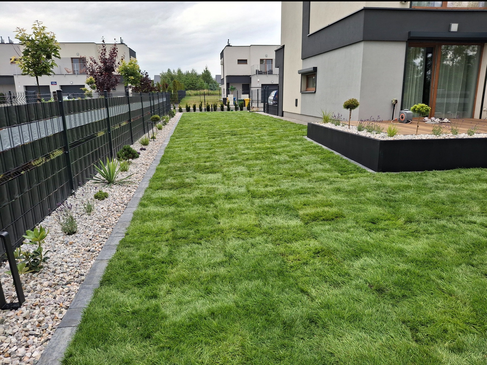 Zadbany trawnik w nowoczesnym ogrodzie z metalowym ogrodzeniem, kamienną rabatą i podwyższoną grządką. Widoczne domy w tle.