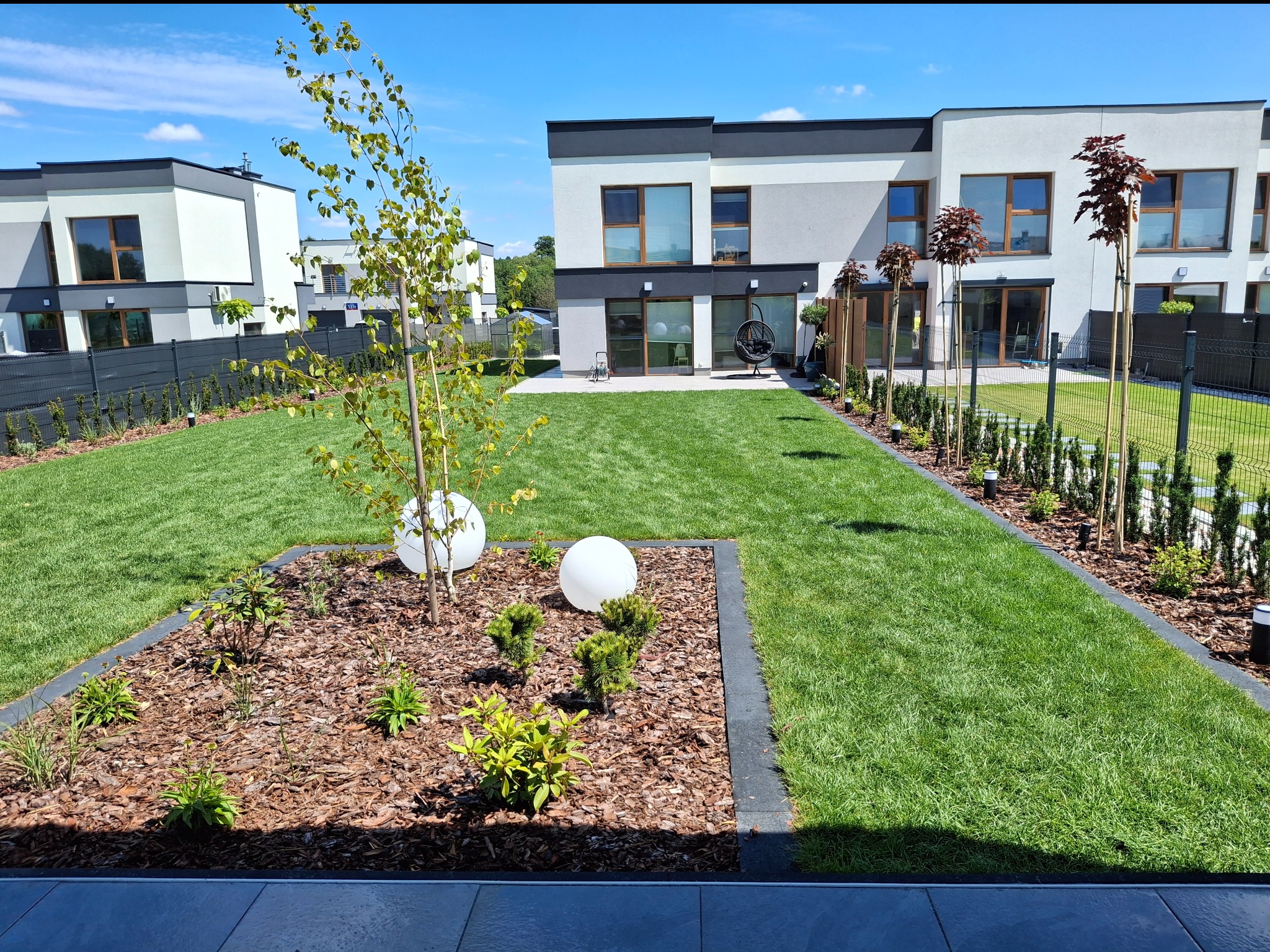 Zadbany trawnik i rabaty z korą w Aleksandrowie Łódzkim. Nowoczesne domy w tle. Regularne koszenie, nasadzenia i oświetlenie podkreślają estetykę ogrodu.