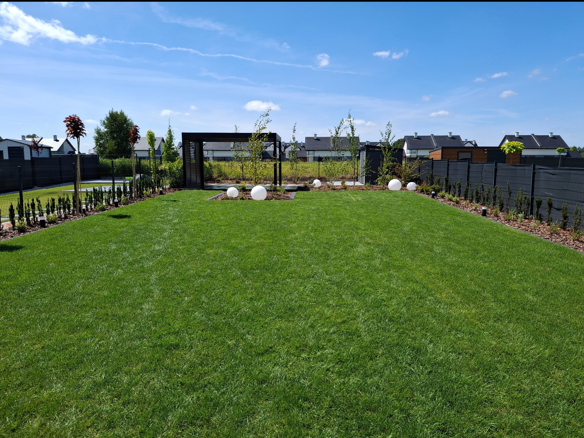 Zadbany trawnik z równo skoszoną trawą, obrzeża z kory, dekoracyjne kule świetlne i pergola w tle, tworzące estetyczny krajobraz przydomowy.