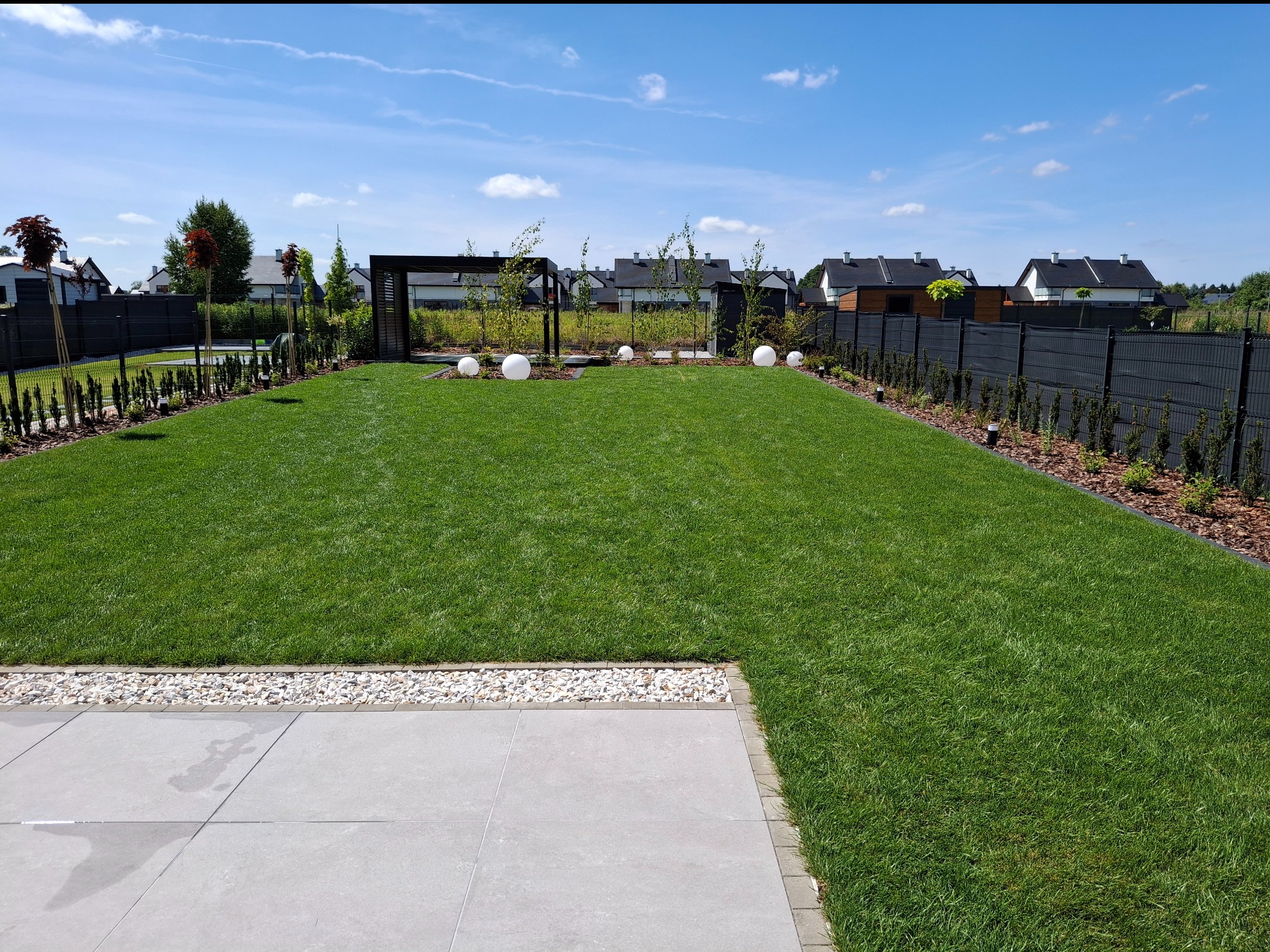 Zadbany trawnik z równo przystrzyżoną trawą, obrzeżami z kamyków i płytami chodnikowymi. W tle nowoczesna pergola i kuliste lampy ogrodowe.