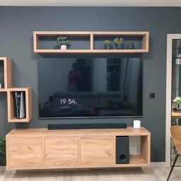 SPIM GRZEGORZ KOCZUR - Nowoczesny salon z szarą ścianą, drewnianą szafką RTV, wiszącymi półkami i telewizorem. Widoczny fragment jadalni z okrągłym stołem i krzesłami. Minimalistyczny design.