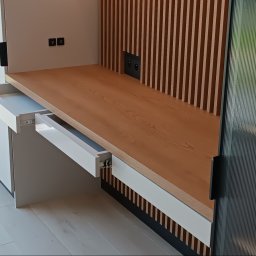 SPIM GRZEGORZ KOCZUR - Nowoczesna zabudowa kuchenna z drewnianym blatem i dekoracyjnymi lamelami. Otwarta szuflada z białym frontem. Widoczne gniazdka elektryczne.