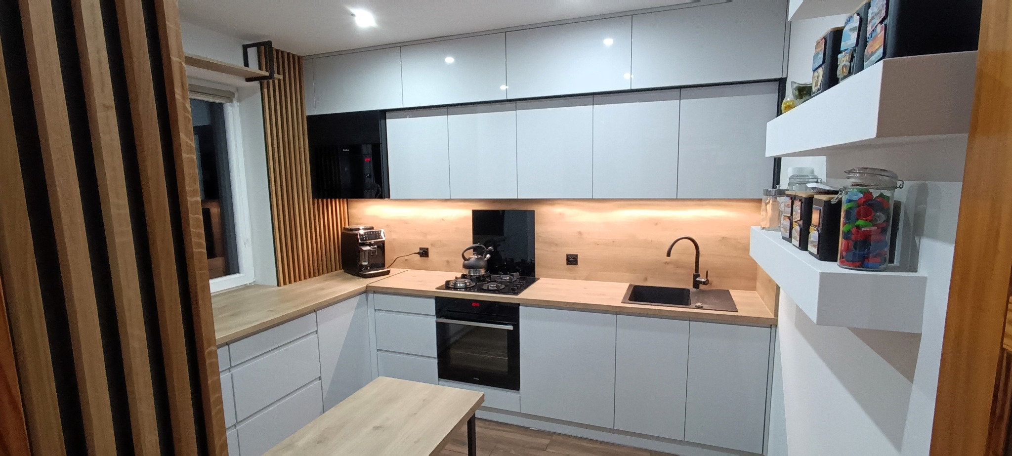 Jasna kuchnia z drewnianymi akcentami: ryflowane panele, blat i półki, ekspres do kawy, czajnik na płycie gazowej.