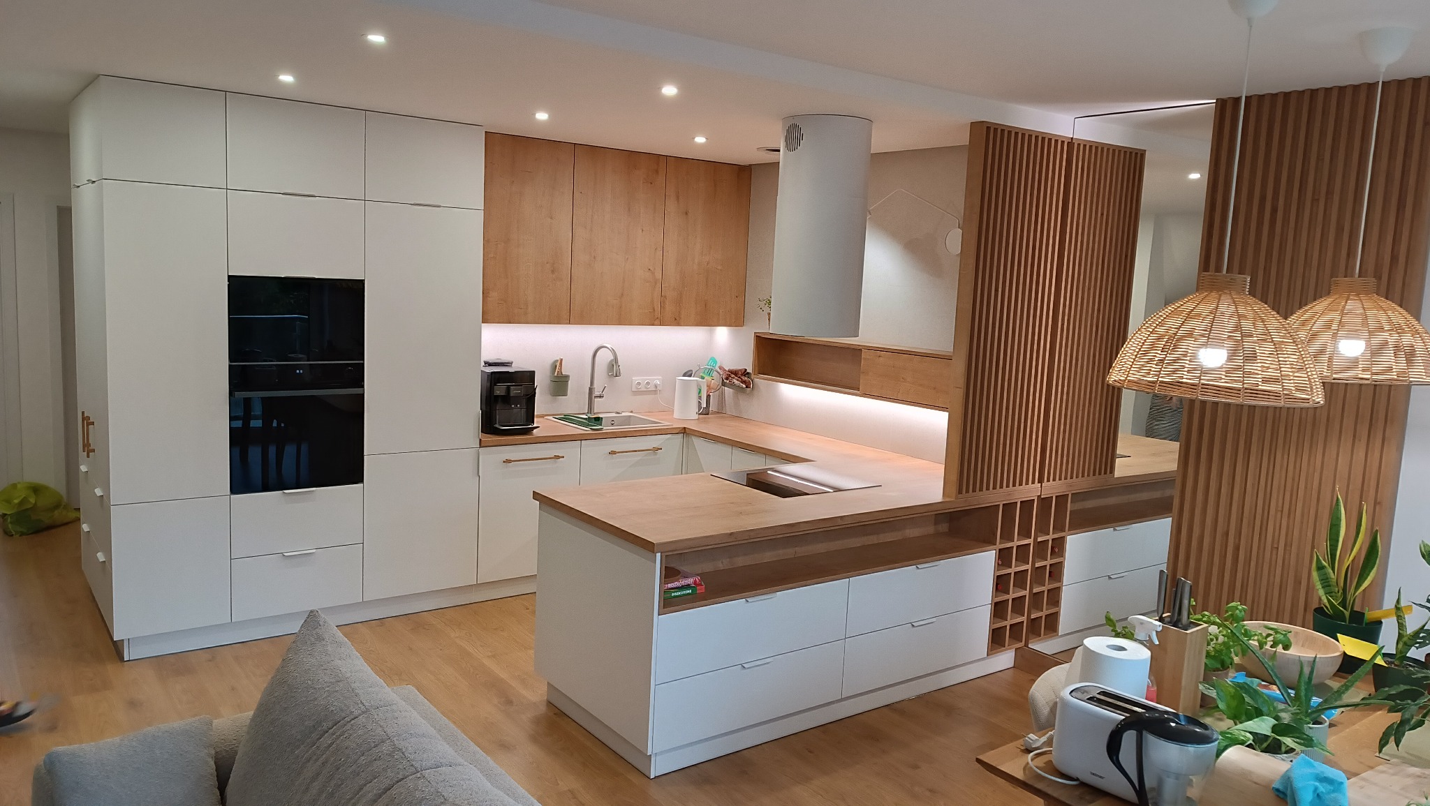 Nowoczesna kuchnia na wymiar z biało-drewnianymi frontami, wyspą i drewnianą ścianką działową. Oświetlenie punktowe i wiszące lampy nadają ciepły klimat.