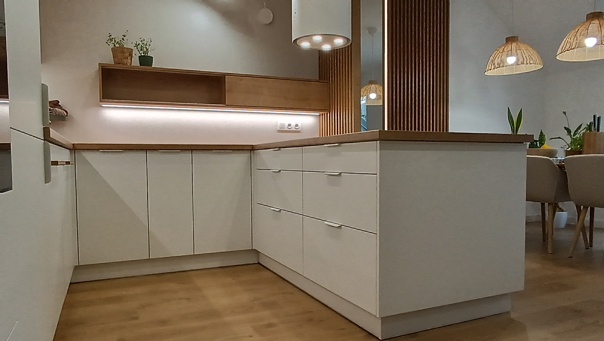 Biała, nowoczesna kuchnia z drewnianym blatem i oświetleniem LED. Widoczna wyspa kuchenna i drewniane lamele dekoracyjne. Uwagę przyciągają plecione lampy.