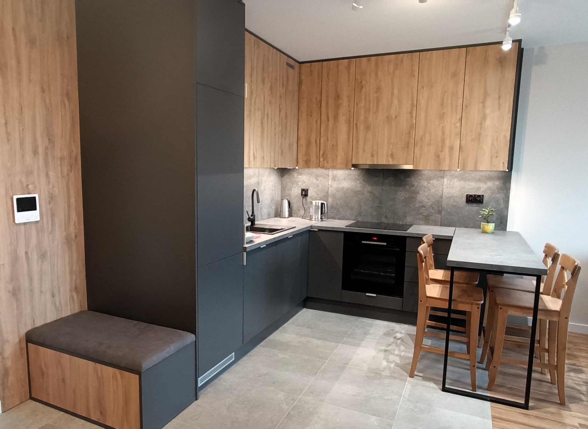 Nowoczesna kuchnia z szarymi szafkami dolnymi i drewnianymi górnymi. Wyjątkowy element to siedzisko z szarą tapicerką wkomponowane w zabudowę.