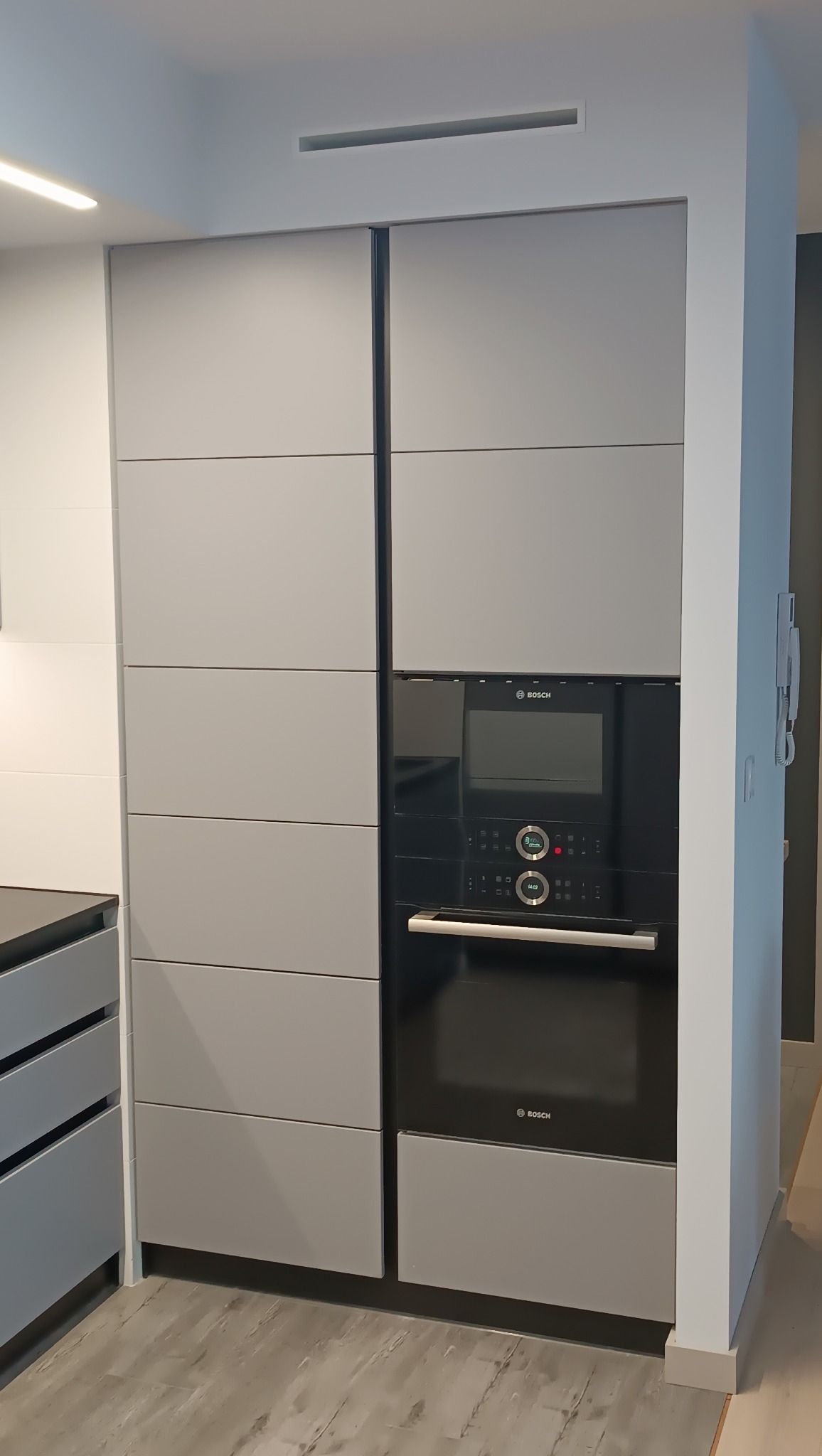 Szara zabudowa kuchenna z piekarnikiem Bosch, minimalistyczny design, poziome podziały frontów, oświetlenie LED podszafkowe, szare panele podłogowe.