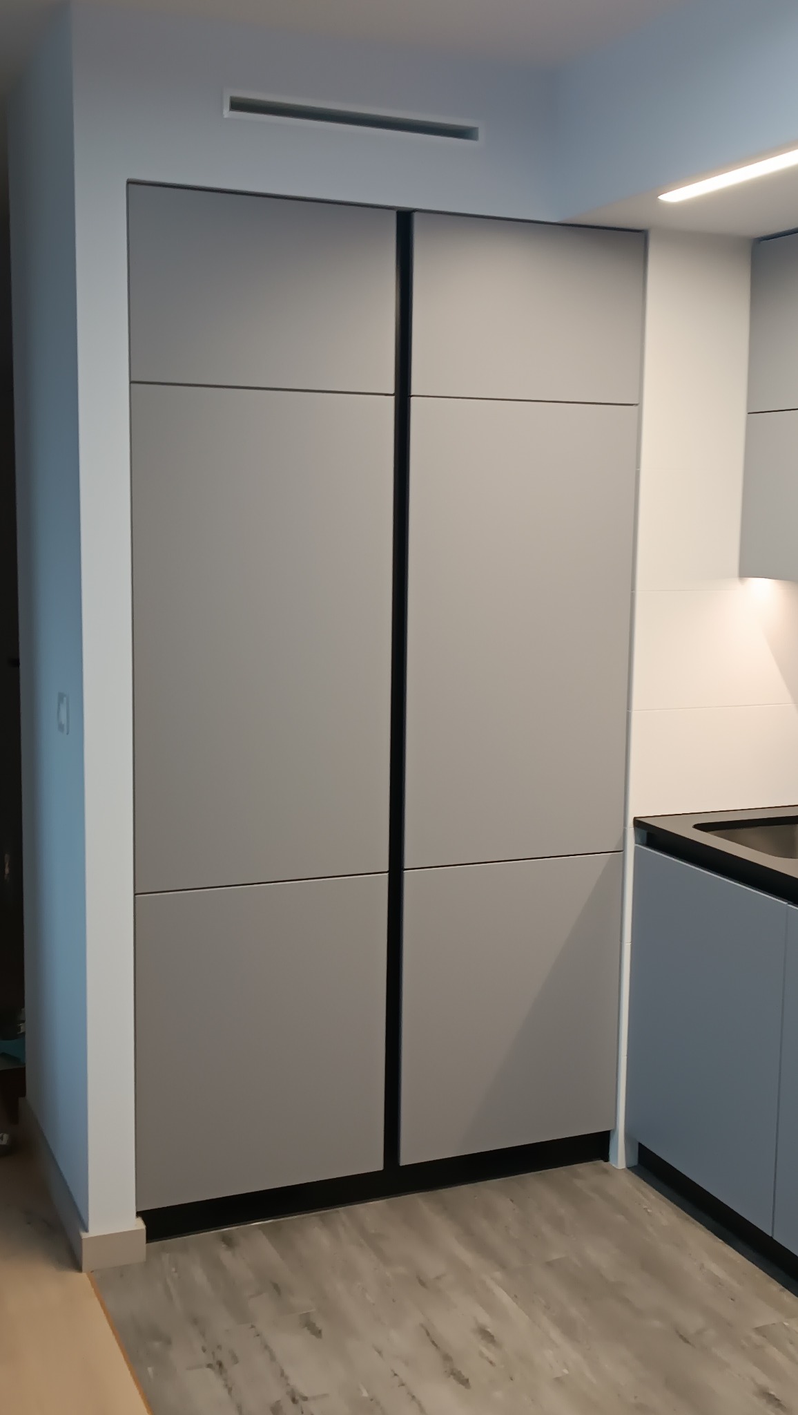 Szare, minimalistyczne szafki kuchenne na wymiar z czterema drzwiami, z czarnymi obramowaniami, na tle drewnopodobnej podłogi i białej ściany z oświetleniem.