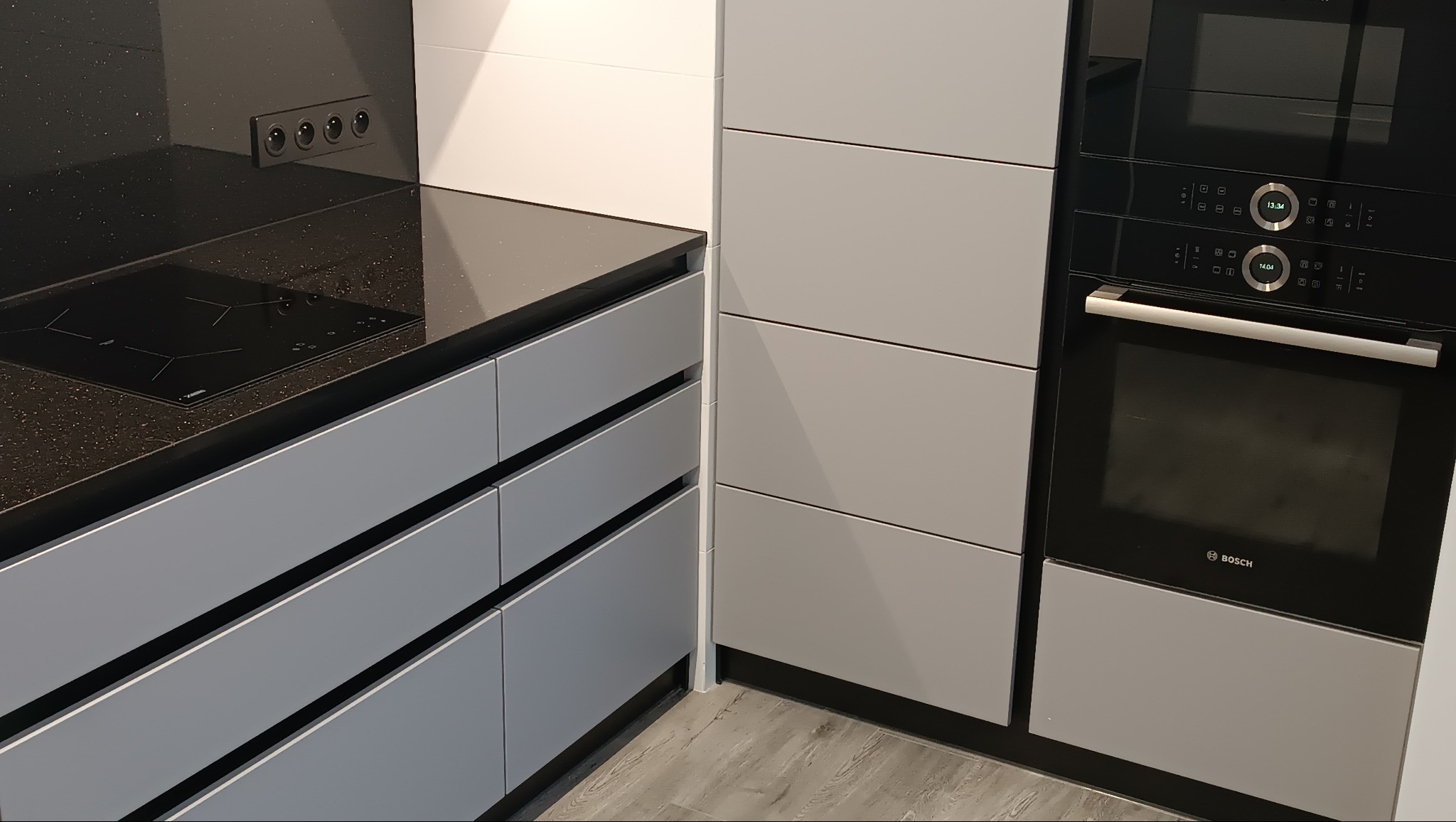 Fragment nowoczesnej kuchni: szare szafki z czarnym blatem, płyta indukcyjna, piekarnik i mikrofalówka Bosch. Minimalistyczny design, gładkie fronty.