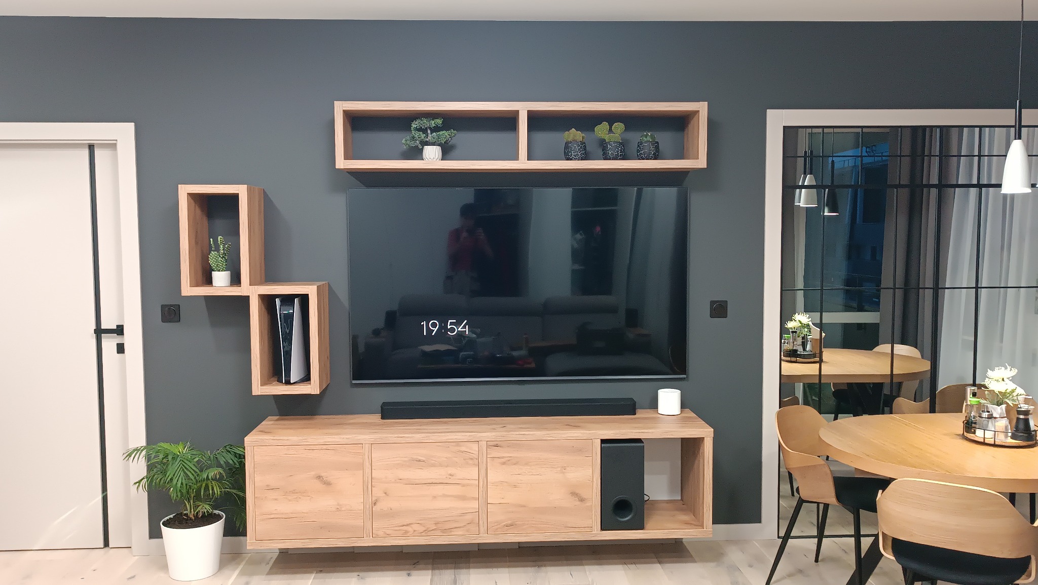 Nowoczesny salon z szarą ścianą, drewnianą szafką RTV, wiszącymi półkami i telewizorem. Widoczny fragment jadalni z okrągłym stołem i krzesłami. Minimalistyczny design.