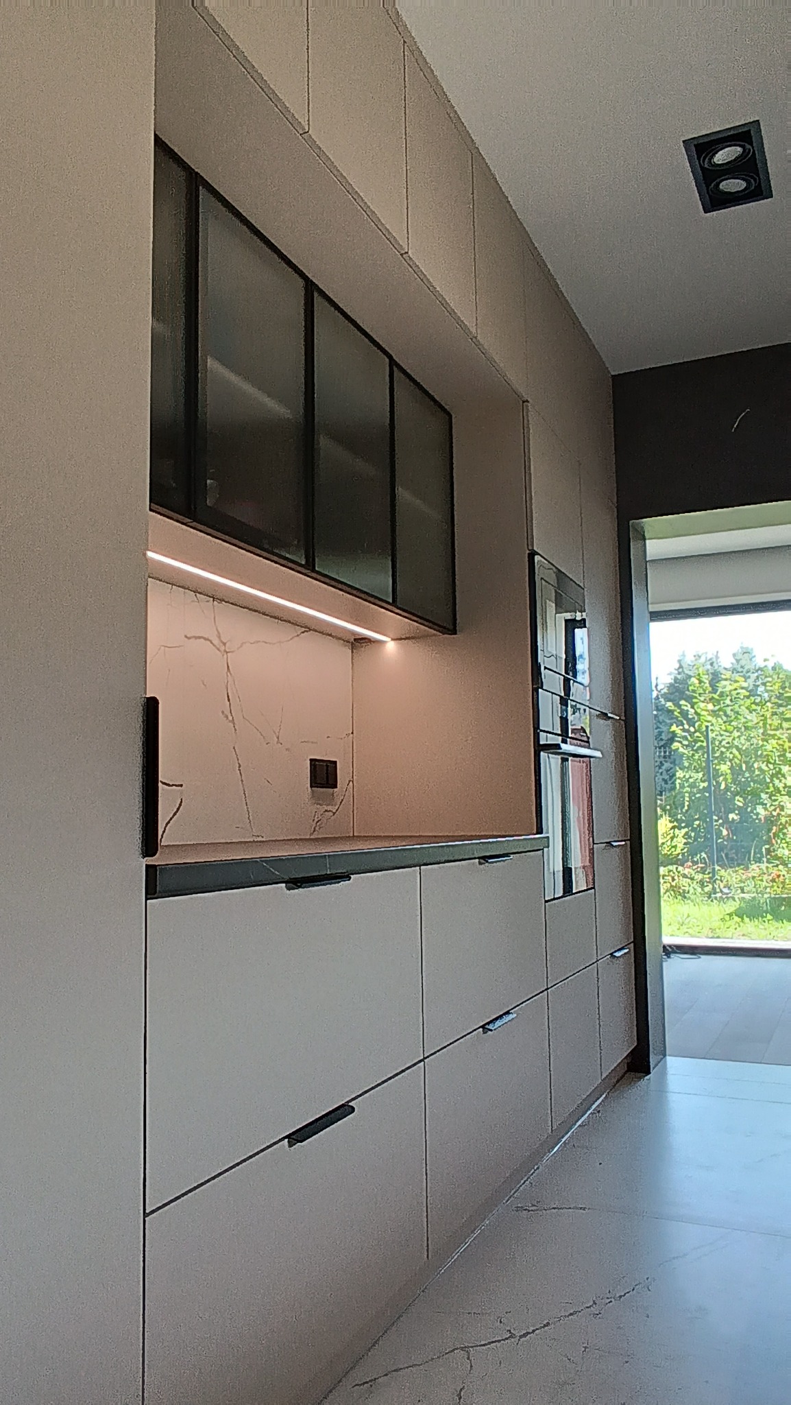 Nowoczesna kuchnia z frontami w kolorze beżowym, czarnymi uchwytami i przeszklonymi szafkami wiszącymi. Widoczne oświetlenie LED i wbudowany piekarnik.