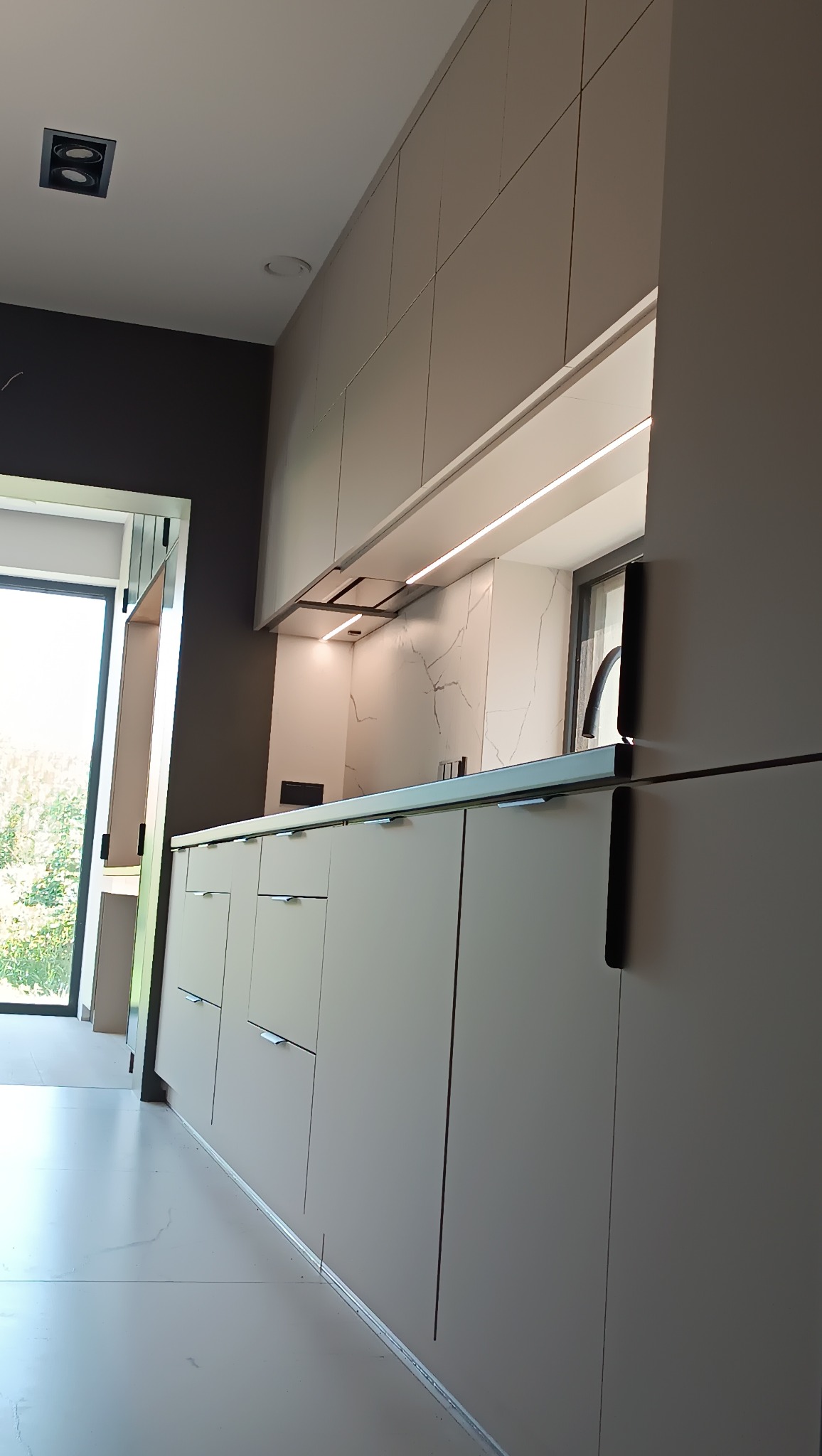 Minimalistyczna kuchnia z beżowymi frontami i marmurowym backsplash'em, oświetlona listwami LED. Widoczne okno z widokiem na zieleń i nowoczesne oświetlenie sufitowe.