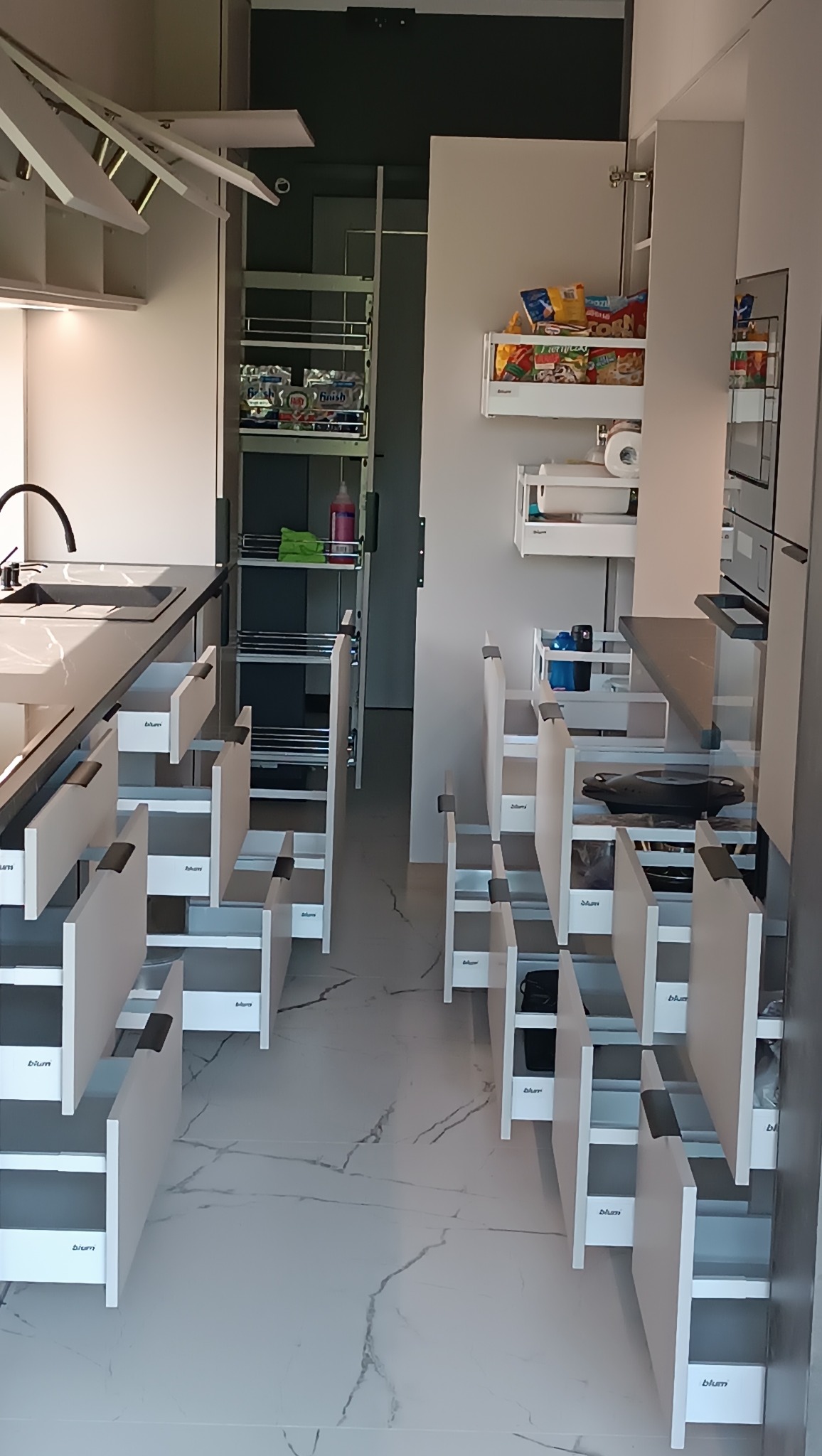 Nowoczesna kuchnia na wymiar z systemem szuflad Blum, otwarte szuflady ukazują funkcjonalne wnętrze. Widoczna spiżarnia i półki z zapasami. Marmurowa podłoga.