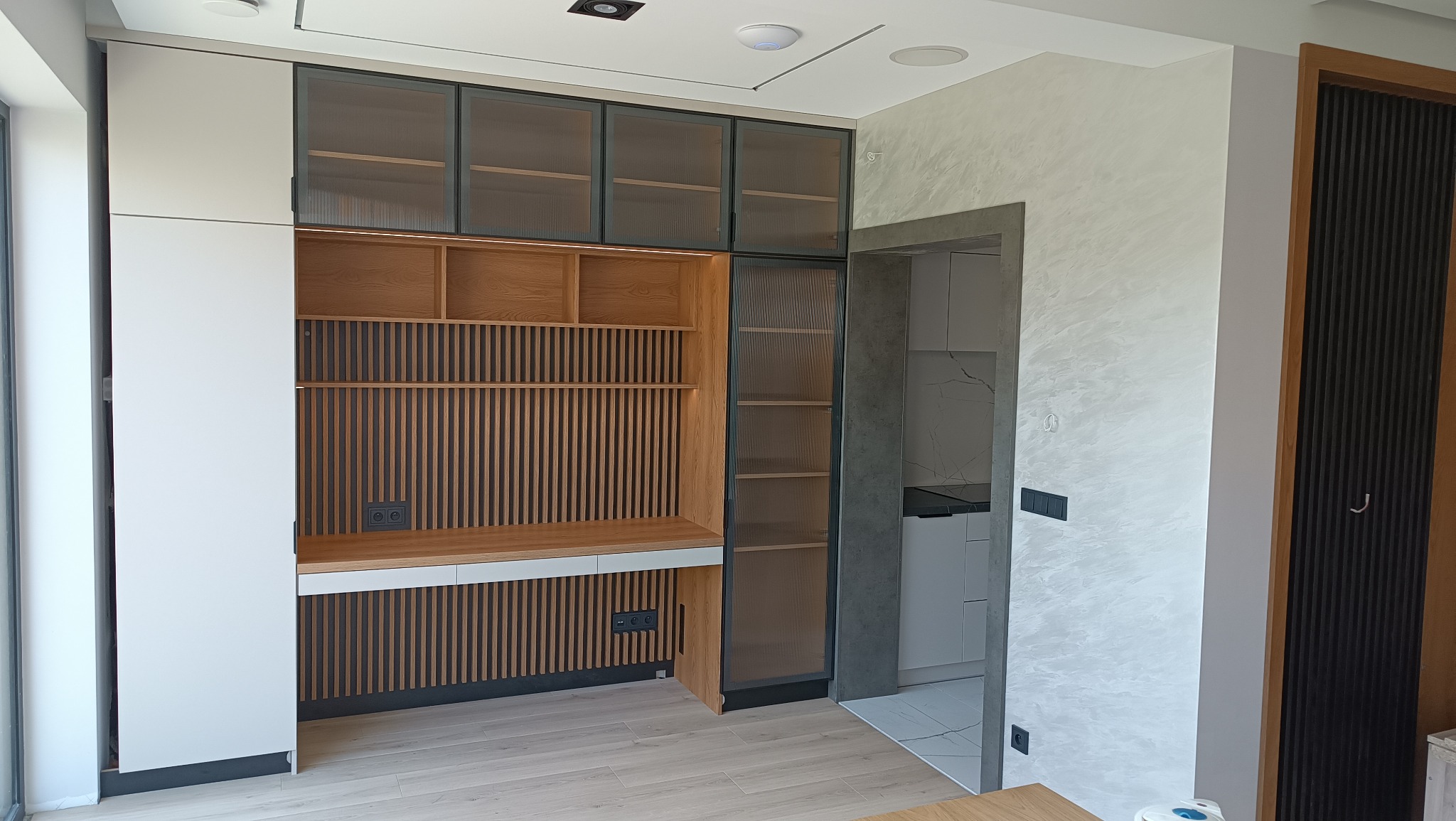 Nowoczesna zabudowa z drewna i szkła ryflowanego, z biurkiem wkomponowanym w strukturę. Widok na wnętrze kuchni przez otwarte drzwi.