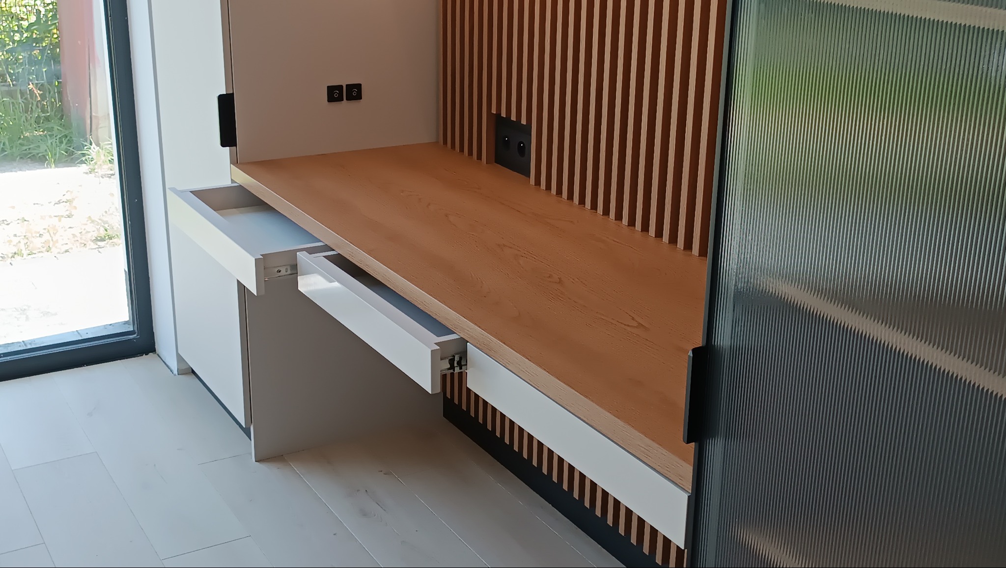 Nowoczesna zabudowa kuchenna z drewnianym blatem i dekoracyjnymi lamelami. Otwarta szuflada z białym frontem. Widoczne gniazdka elektryczne.