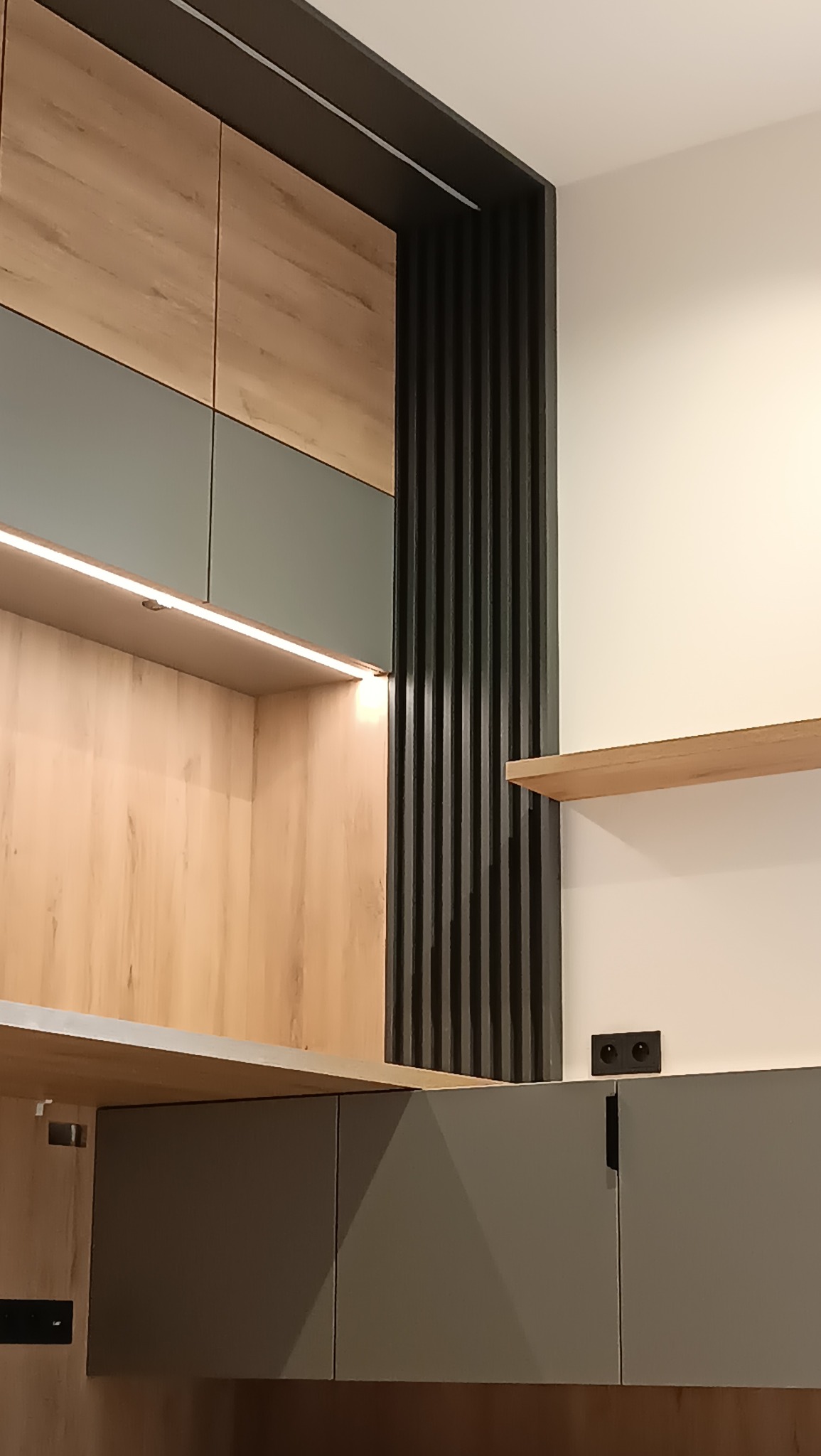 Szare i drewniane szafki kuchenne z oświetleniem LED i czarnymi dekoracyjnymi listwami. Widoczny fragment blatu i gniazdko elektryczne.