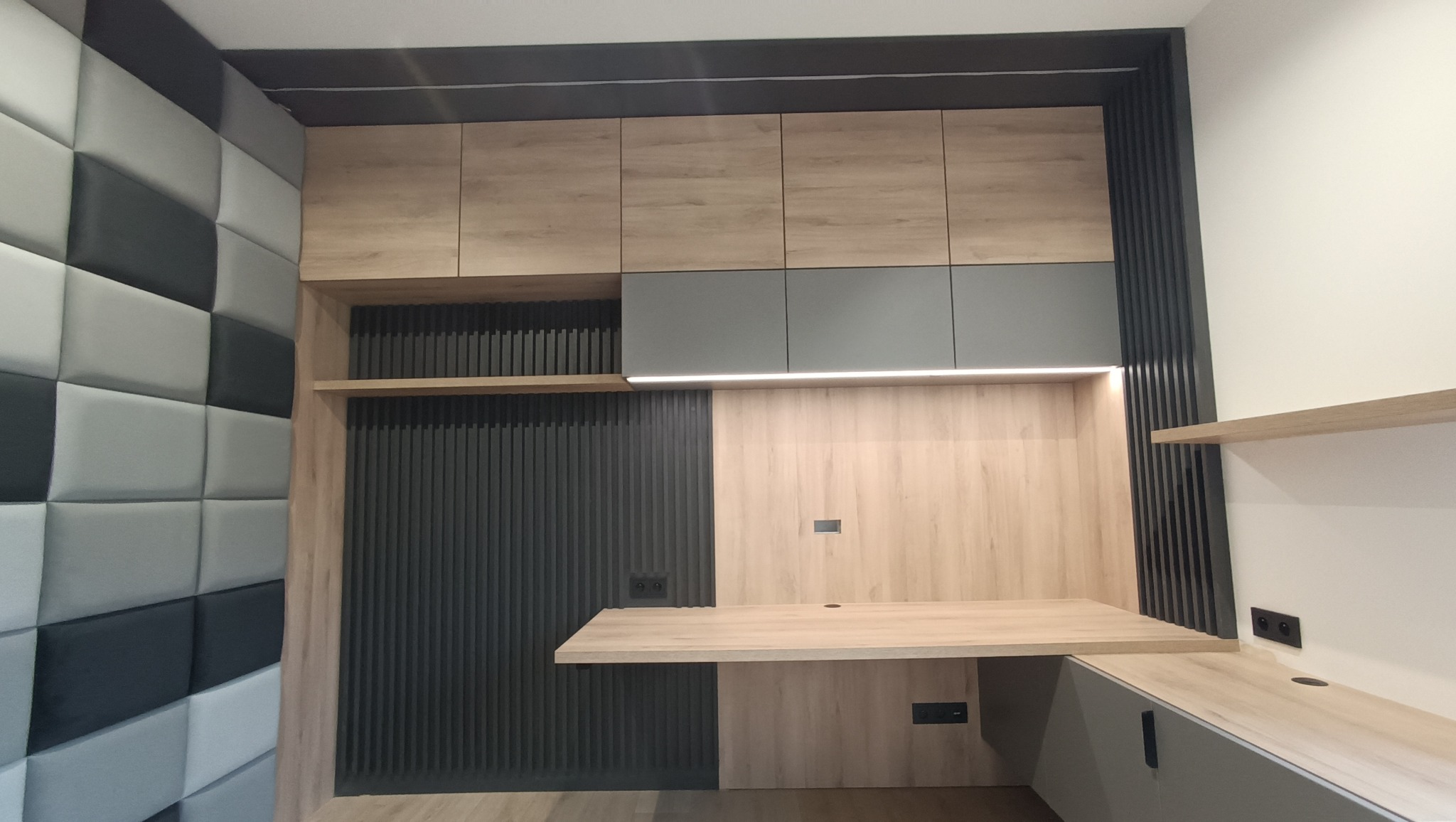 Nowoczesna zabudowa kuchenna z drewnianymi szafkami, szarymi akcentami i podświetleniem LED, uzupełniona o panel ścienny w geometryczny wzór.