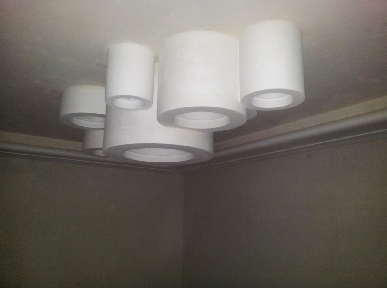 Nietypowa lampa sufitowa w kształcie białych, grubych rur o okrągłym przekroju, zamontowana na białym suficie w rogu pokoju o szarych ścianach.