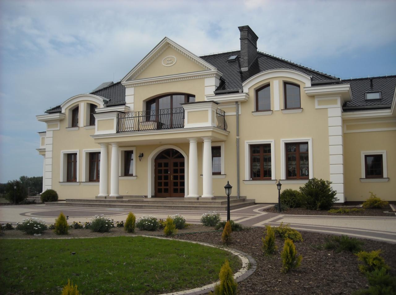 Elegancki, dwukondygnacyjny dom z balkonem, kolumnami i ciemną dachówką, otoczony starannie utrzymanym ogrodem.