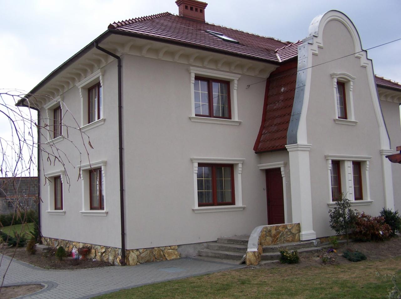 Elewacja domu jednorodzinnego z jasnym tynkiem, czerwoną dachówką, kamienną podmurówką i ozdobnymi gzymsami okiennymi.