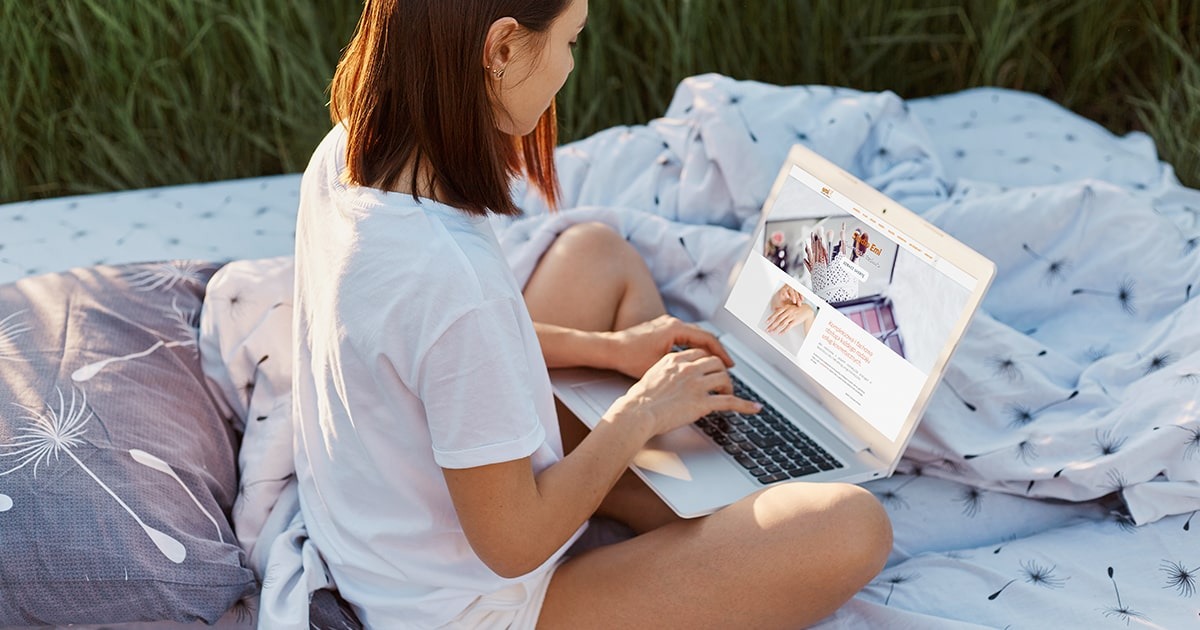 Kobieta w białej koszulce siedzi na kocu w trawie i pracuje na laptopie, na ekranie widoczna strona internetowa z prezentacją kosmetyków i usług kosmetycznych