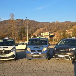 Trzy busy taxi r&oacute;żnych marek (Renault, Toyota) stojące na parkingu z widokiem na g&oacute;rzysty krajobraz, w słoneczny dzień. Tablice rejestracyjne widoczne.