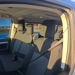 F.H.U. Dunajec Zbigniew Surowiak - Wnętrze busa z widocznymi fotelami pasażerów, pasami bezpieczeństwa i uchwytami. Z okna widać zabudowania miejskie. Oświetlenie słoneczne podkreśla detale.