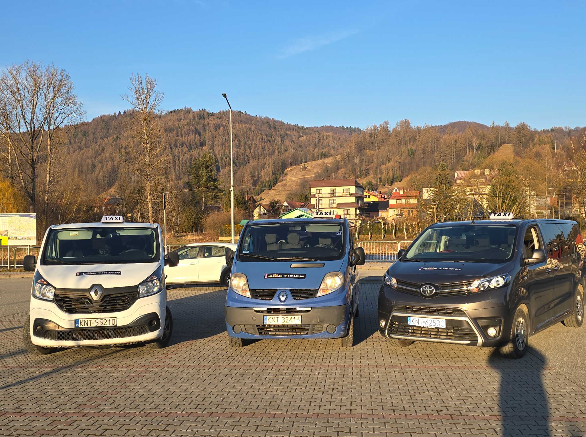 Trzy busy taxi różnych marek (Renault, Toyota) stojące na parkingu z widokiem na górzysty krajobraz, w słoneczny dzień. Tablice rejestracyjne widoczne.