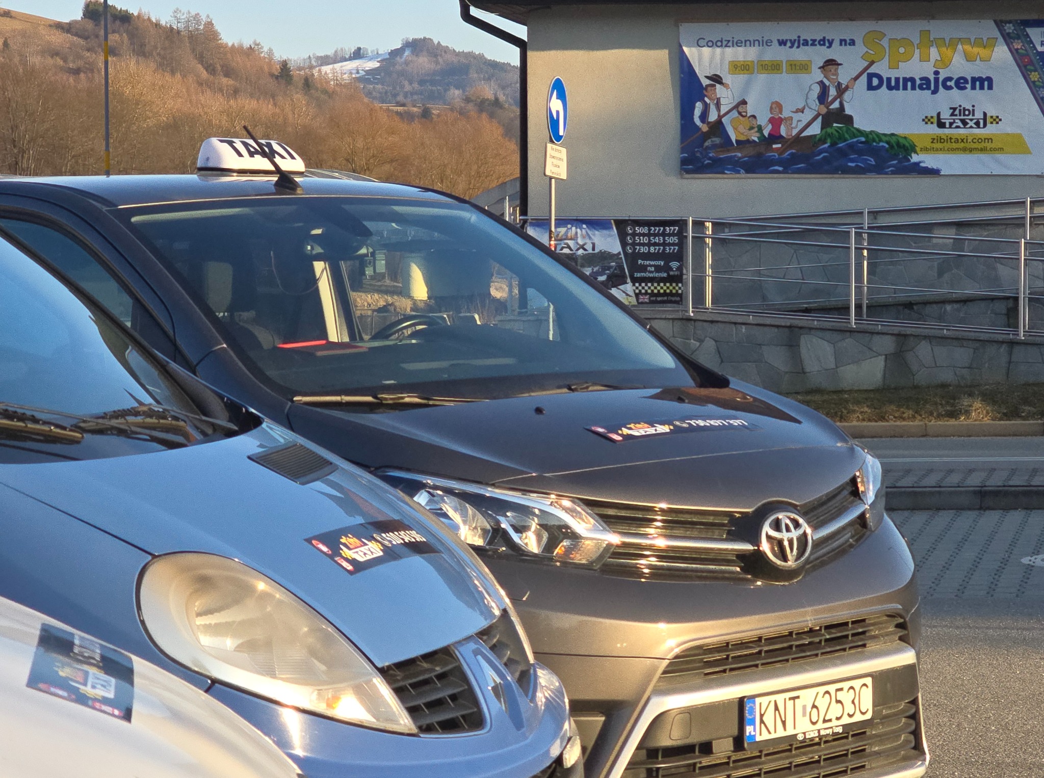 Dwa auta taxi zaparkowane obok siebie. Na budynkach reklamy spływu Dunajcem. Szaro-niebieski i grafitowy samochód z napisem TAXI na dachu i naklejkami na karoserii.