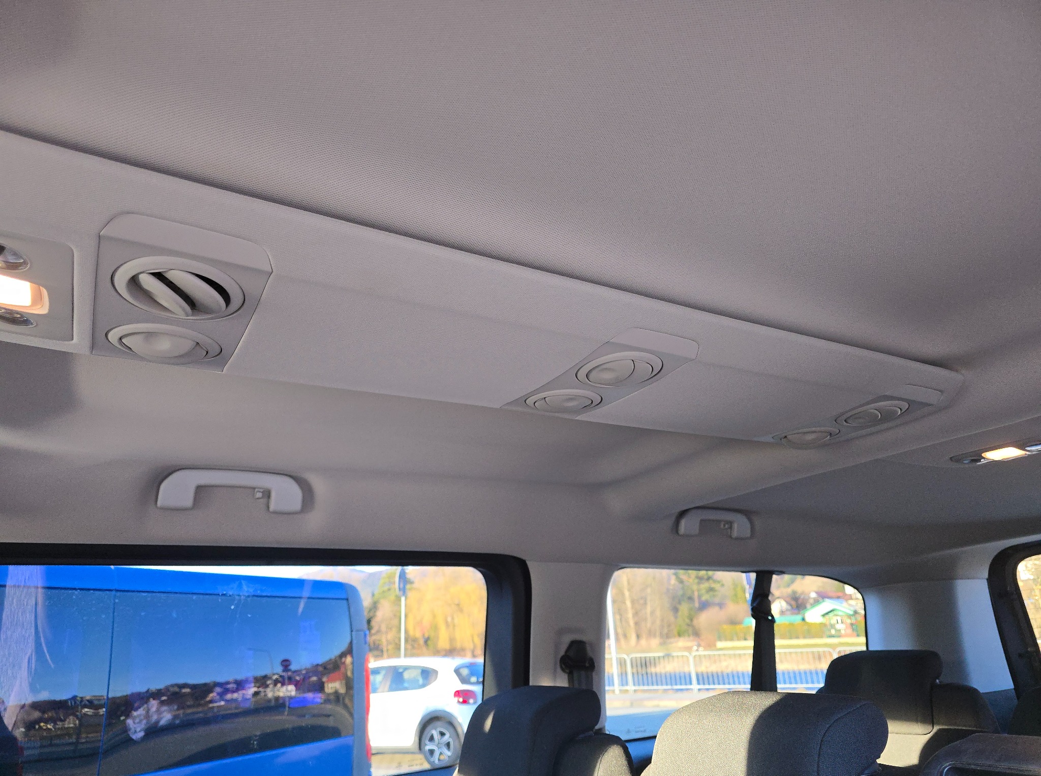 Wnętrze pojazdu z widocznymi dyszami wentylacyjnymi w suficie, uchwytami dla pasażerów i oknami z widokiem na zewnątrz. Widoczne fotele i elementy konstrukcyjne pojazdu.