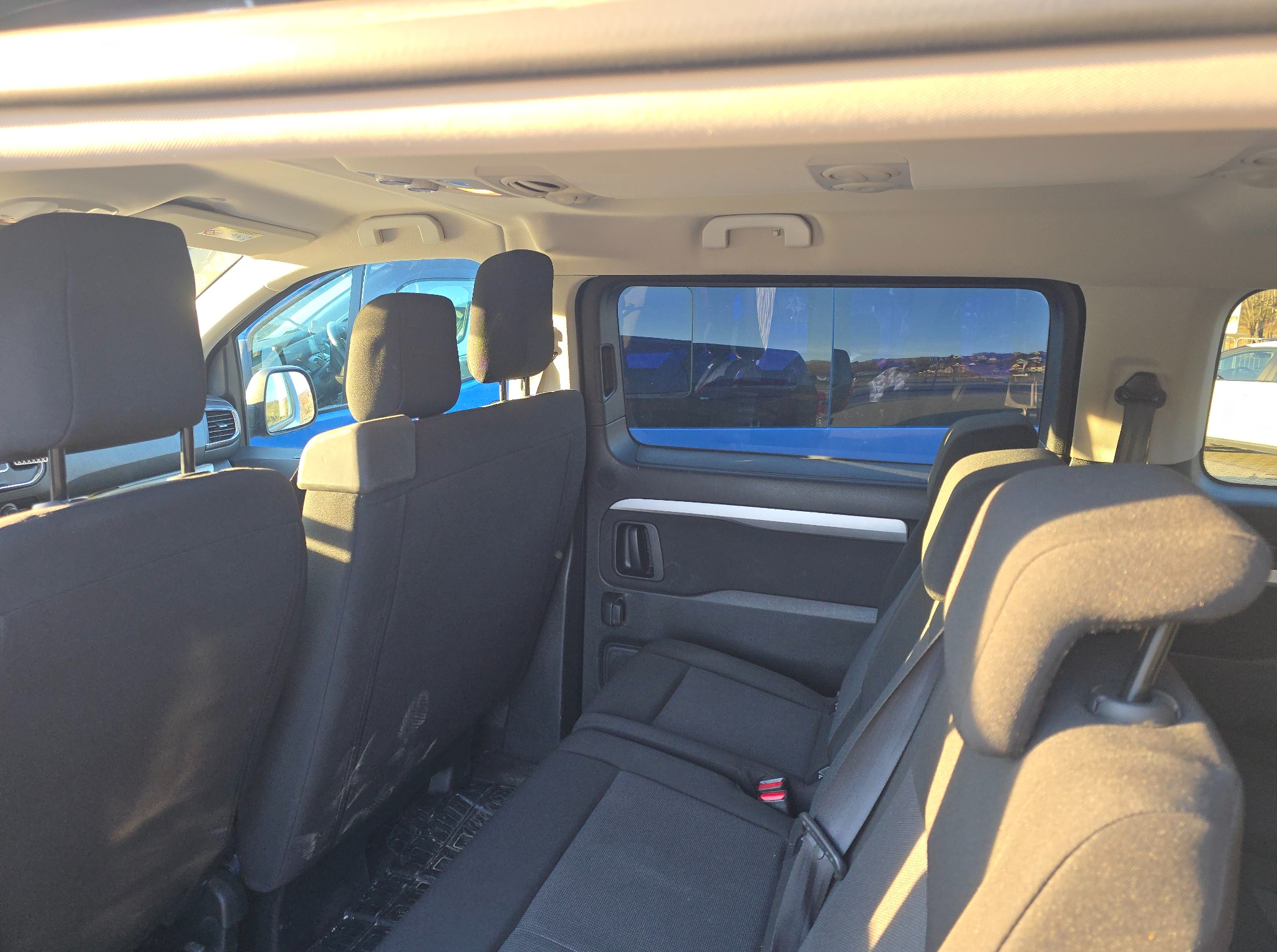 Wnętrze busa z trzema rzędami czarnych foteli, widok z tyłu pojazdu. Przez okno widać inne pojazdy i fragment krajobrazu. Oświetlenie słoneczne.